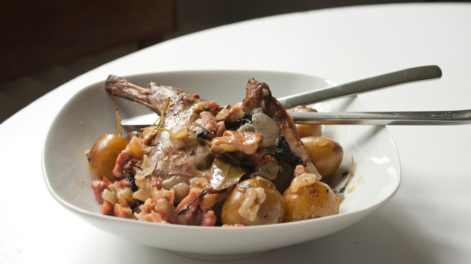 Assiette blanche pos&eacute;e sur une table et contenant du lapin &agrave; la moutarde et deux couverts