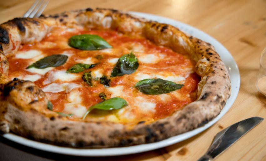 ALT P i zza Napolitaine sur une assiette blanche p osée sur une table en bois et entourée d une fourchette à gauche et d un couteau à dr oite
